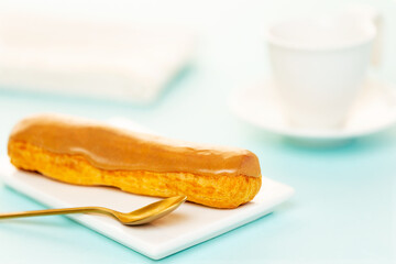 Salty caramel French eclair pastry on white plate, aside a golden spoon and a white coffee cup over light blue background.