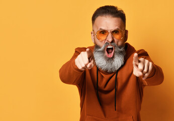 Brutal fed-up grey-haired bearded hipster man in sportswear and trendy sunglasses shouting or yelling loudly and angrily expressing accusation. Sport fashion, people emotion. Studio shot on yellow