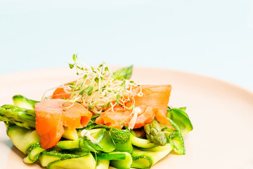 Smoked salmon salad with bean sprout, asparagus, enoki mushrooms, zucchini and beetroot on a pink plate over a blue background.