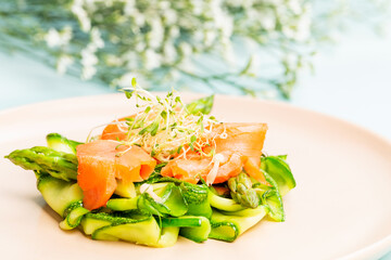 Smoked salmon salad with bean sprout, asparagus, enoki mushrooms, zucchini and beetroot on a pink plate over on a blue background with twigs of white flowers.