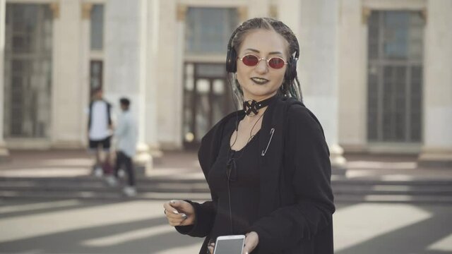 Carefree Caucasian hippie woman in sunglasses and headphones dancing on sunny city street with blurred men talking at background. Portrait of relaxed young goth enjoying music outdoors.