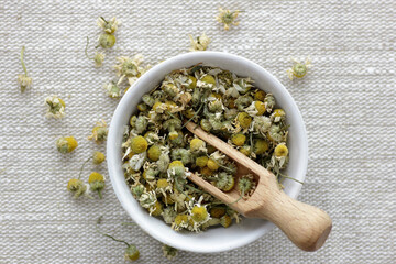 Chamomile dried flower tea in a white bowl on linen textile with blossoms and buds nearby, closeup, copy space, flat lay, from above overhead top view, healthy herbal teas and natural healer concept