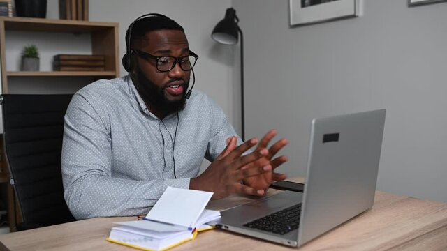 Side view of an African guy enthusiastically talking online using hands free headset for it. A multiracial young man has online conversation, conducts webinar sitting at home office