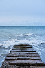 Fototapeta premium Waves break on rocks and fly apart into small drops and turn into foam. Long concrete breakwater stretching away into depths of stormy blue sea.