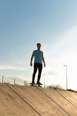 Young man wearing a blue t-shirt and black trousers riding a skateboard with a blue sky