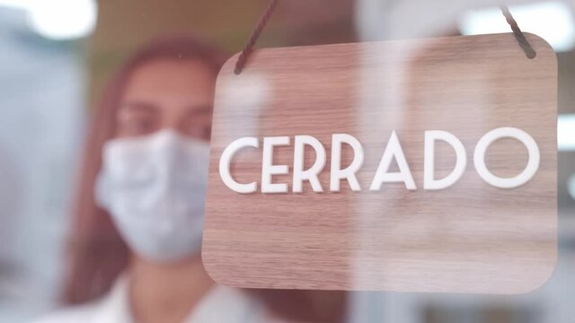 Young sad woman with face mask changing open to closed sign in spanish on window for corona virus pandemic. 4k 60fps