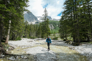 A backpacker man standing by a rushing torrent in region of Dachstein, Austria. There is dense forest behind, and high Alpine peaks. Taking a moment to relax. Admiring the beauty of the nature.