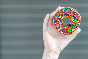 Hand wearing latex gloves, holding a fresh chocolate frosted donut and looking at the hole of the donut. Playful and joyful tasty sugary comfort food for customers.