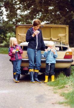 Vintage 1977 image of a young mother with son and daughter standing and eating a sandwich in nature, in front of a seventies Ford Taunis with open car trunk.	
