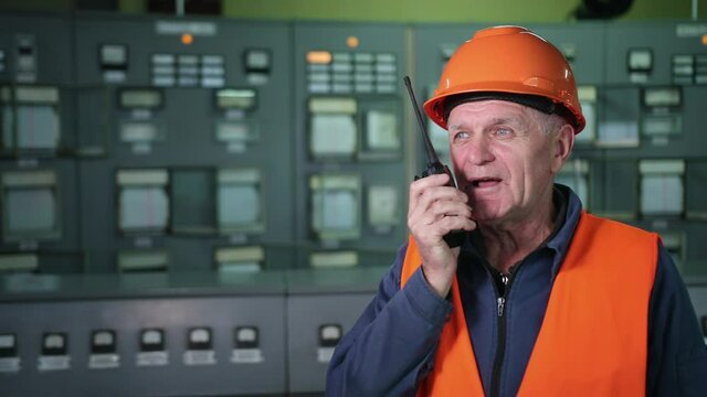 Check control. Male engineer in a orange helmet looks at the control panel with buttons indication, speaks on walkie-talkie. Close up engineer man wearing face mask, using walkie-talkie in factory.