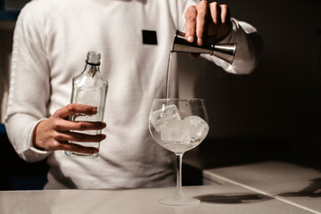 Bartender pouring a white transparent alcoholic drink from the steel jigger to a cup with ice cubes on the bar counter