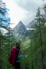 Naklejka premium A backpacker woman in pink jacket pointing to the peak of Marstein, Austrian Alps. The slopes are partially covered with snow. Stony and sharp mountains. Overcast. Dense forest at the foothill. Happy