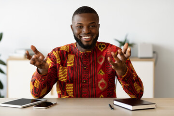 Smiling african man sitting at desk in home office, talking at camera