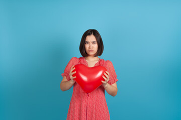 close up portrait of a sad upset disappointed young Asian woman holding a red heart shaped balloon on Valentine's day isolated on a blue background.