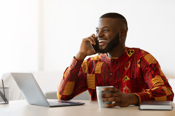 African Freelancer Guy Talking On Cellphone And Enjoying Coffee At Home Office