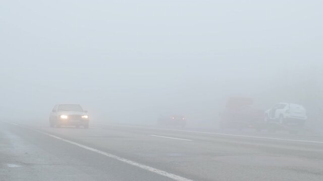 Car drives in fog along road in autumn, at daytime. Very poor visibility on track due to thick mist, side view. Passenger car drives with headlights in dangerous haze covered highway. Foggy weather