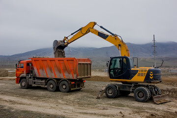 Excavator at a construction site loads a truck against a background of mountains