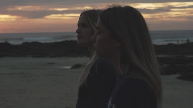 Two silhouetted young girls walk in slow motion down a beach past a golden sunset with a rocky coastline in the background.
