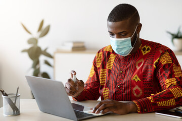 Hygiene On Workplace. African Man In Mask Sanitizing Laptop Keyboard With Disinfectant