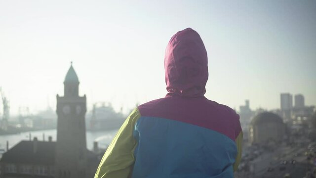 young girl or women puts on hood in hamburg city, girl wearing colorful rain jacket and standing back to the camera, sun is shing on cloudy day during coronavirus lockdown.