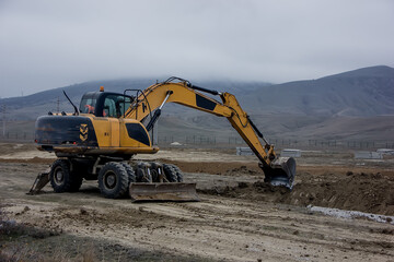 Obraz premium Yellow excavator digs a trench against the backdrop of mountains.