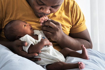 An African American father kissing hand,  his 12-day-old baby newborn son lying in bed in a white...