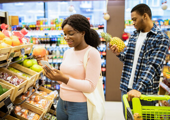 Lovely black woman and her boyfriend shopping for fruits, checking grocery list on mobile phone at...