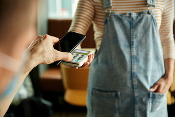 Woman wearing face mask behind cafe counter holding contactless payment device while customer uses mobile phone to pay bill