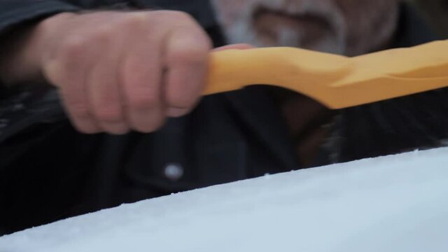 Aged businessman with grey beard cleans electric car roof with black and yellow snow brush on cold day. Concept harsh weather