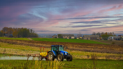 Niebieski traktor rozpyla nawóz, w tle majestatyczne niebo i łąka. 