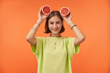 Teenage girl, cheerful and happy with short brunette hair holding grapefruit over her head. Standing over orange background. Wearing green t-shirt, teeth braces and bracelets