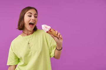 Portrait of attractive, nice looking girl holding an ice cream and looking at the right to the copy space over purple background. Wearing green t-shirt, bracelets, rings and necklace