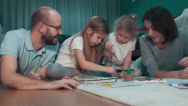 
Mother and father teach their two daughters draw pictures in the living room on the floor.