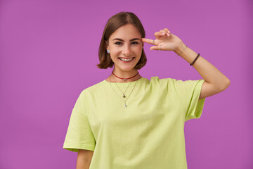 Teenage girl, cheerful and happy, with brunette short hair. Holding a finger next to her temple. Standing over purple background. Wearing green t-shirt, teeth braces, necklace, bracelets and rings