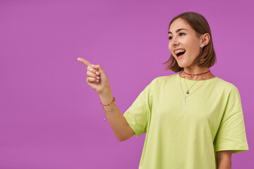 Female student, young lady, laughing and pointing her finger to the left at the copy space over purple background. Showing a sign. Wearing green t-shirt, teeth braces, bracelets and rings