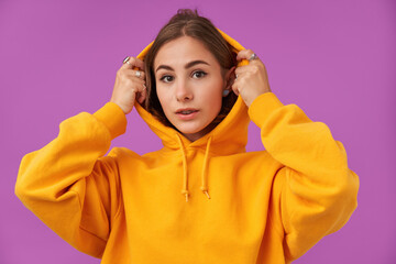 Fototapeta na wymiar Portrait of attractive, nice looking girl with brunette short hair. Putting hood on and watching at the camera over purple background. Wearing orange hoodie, braces and rings