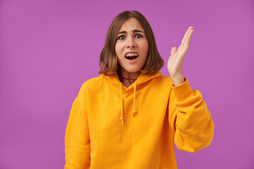 Portrait of attractive, nice looking girl with short brunette hair, shocked, horrified lady lifts her hand. Looking at the camera over purple background. Wearing orange hoodie, teeth braces and rings