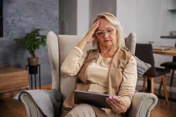 Blond senior woman sitting at home, having headache and looking at tablet to get online advices.