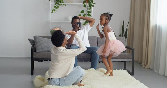 Three people young afro american family sitting in living room interior father takes phone camera takes photo while little daughter jumps from couch to floor on carpet gives mom five symbol of support