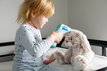 Smiling little child measures temperature of toy fluffy bunny with non-contact thermometer. Cute small girl sitting on the bed, playing doctor with her stuffed toy
