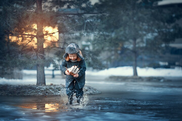 Cute funny little boy in cap with ear-flaps playing with toy little ship and puddles in yearly spring during the sunset in Russia. Image with toning and selective focus