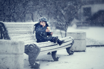 Portrait of russian cute funny little boy in cap with ear-flaps eating a piece of bread sitting on the snowy bench in snowy winter park during the snowfall. Image with toning and selective focus