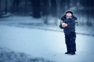 Fototapeta na wymiar Cute little boy in hat with ear-flaps with piece of bread looking up on falling snow in winter park in Russia. Image with toning and selective focus