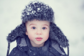 Portrait of russian cute funny little boy in cap with ear-flaps in snowy winter park during the snowfall. Image with toning and selective focus