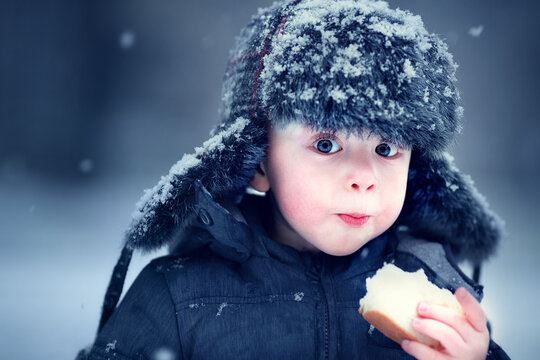 Portrait of russian cute funny little boy in cap with ear-flaps with piece of bread in snowy winter park during the snowfall. Image with toning and selective focus