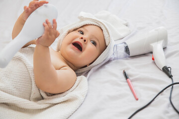 beautiful baby with a white towel on her head lies near the hairdryer, cosmetics and holding a comb