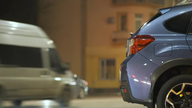 Blue car parked on brightly illuminated city street with moving traffic lights on background at night.