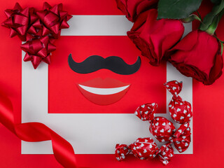 
White frame, candy, ribbon, roses and a smile with a mustache on a red background, close-up top view.