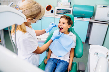 A small patient with an open mouth at a dentist appointment in a modern clinic. Hands of unrecognizable pediatric dentist doing examination procedure for smiling cute boy in hospital
