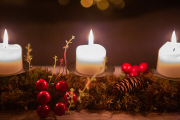 Three burning candles on a dark background with red berries, moss and pine cone 
